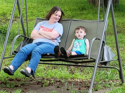 A picture of a woman wearing a gray shirt and blue jeans and child wearing a green shirt and blue shorts on a swing.