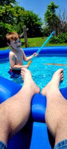 Vertical photo of a shirtless boy in a small swimming pool with adult legs in the foreground.