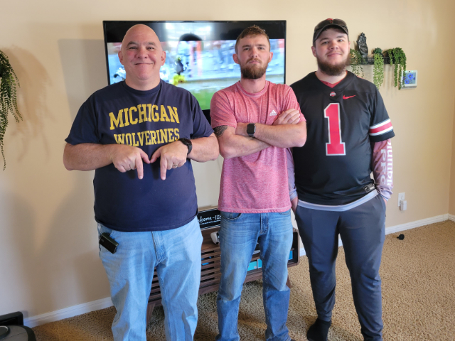Three men standing in front of a TV. The man on the left is wearing a blue Michigan Wolverines t-shirt and making an M with his hands. The man in the middle is wearing a red shirt and has a sour expression. The man on the right is wearing a black Ohio State jersey. | Thanksgiving Joy, Gridiron Drama, and Neighborly Love
