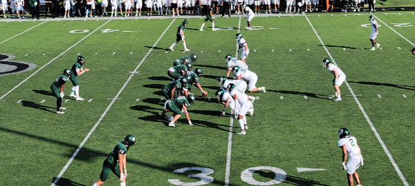 Image of the line of scrimmage at a college football game. Northeastern State is on offense on the left wearing green jerseys. Northern Missouri State is on defense on the right wearing white jerseys.