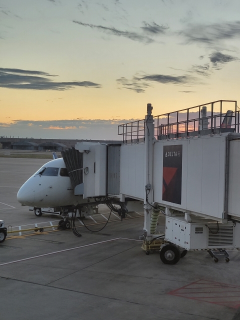 Image of the front of an aircraft at an airport gate. The jet bridge is extended from the right and the sun is just rising behind the jet bridge.
