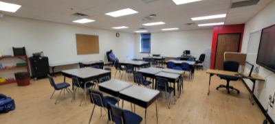 A wide shot of a school classroom with desks and chairs arranged in clusters. A woman in a blue shirt sits in a chair in the background. | Science Under Scrutiny – A Closer Look At Junk Science