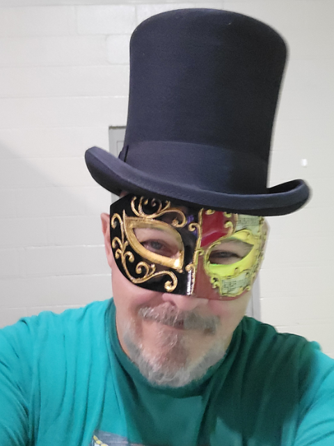 Image of a bearded man wearing a black top hat and red and black Mard Gras mask. He's wearing a green shirt.