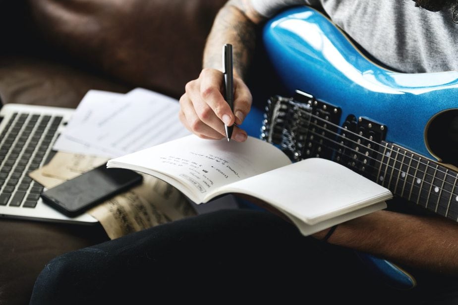 Caucasian man holding a blue guitar while writing song lyrics in a notebook. | When the Lyrics Hit Home: Songs I’ll Never Forget