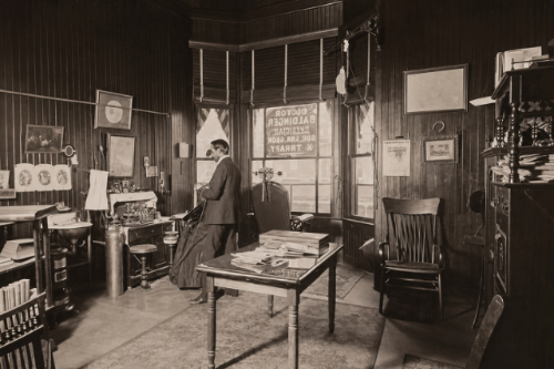 B&W image of a man standing near the back wall of his Eye, Ear, Nose and Throat practice. He's facing the wall and standing in front of a table with medical equipment on it. There's a table in the foreground with books and papers. In the background, there's a large picture window, and in front of that is his examination chair.