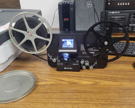 Image shows a digitizer for home movies with a full reel on the left and an empty one on the right. Other computer equipment is visible in the background.